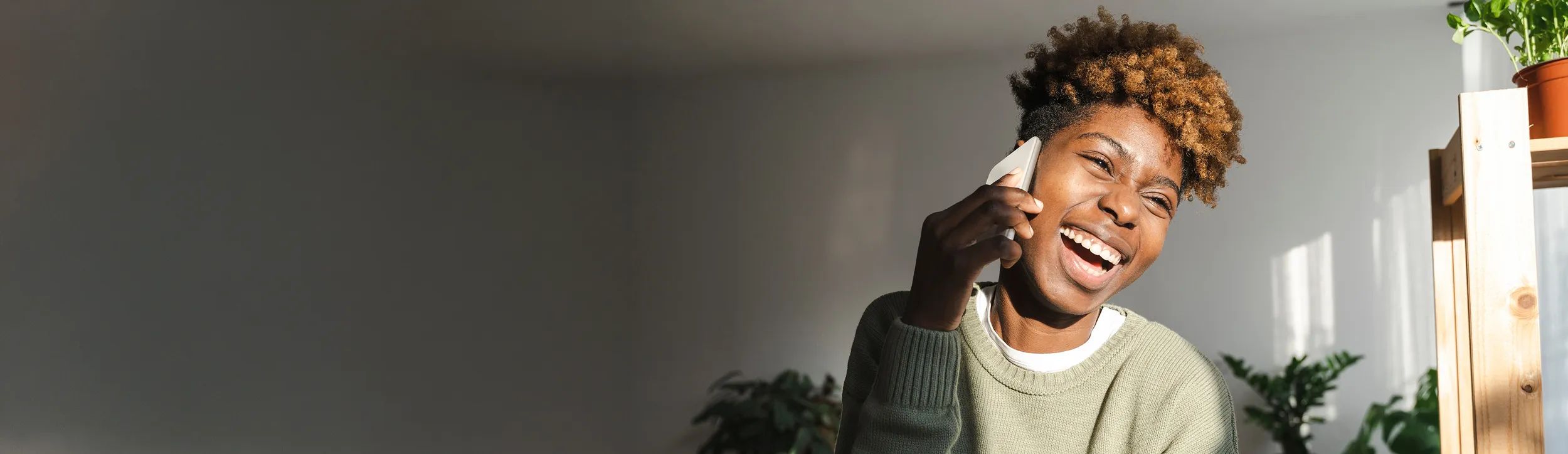 Een jonge vrouw glimlacht terwijl ze met haar mobiel apparaat praat in een lichte, moderne woning met planten.